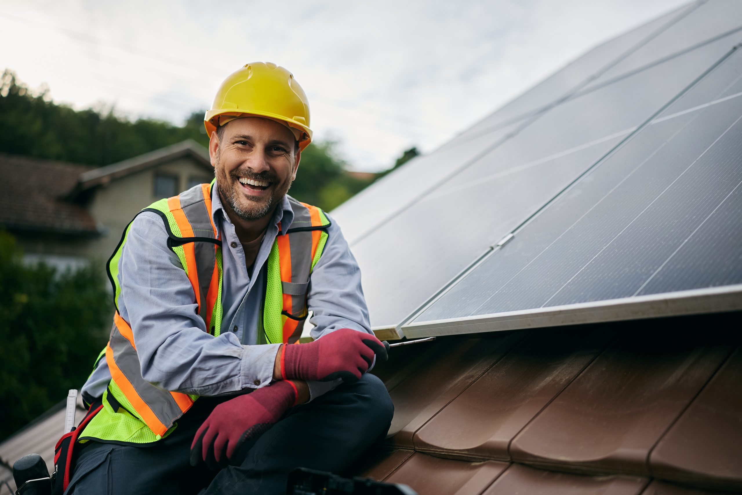 Smiling solar technician kneeling on a residential rooftop beside solar panels