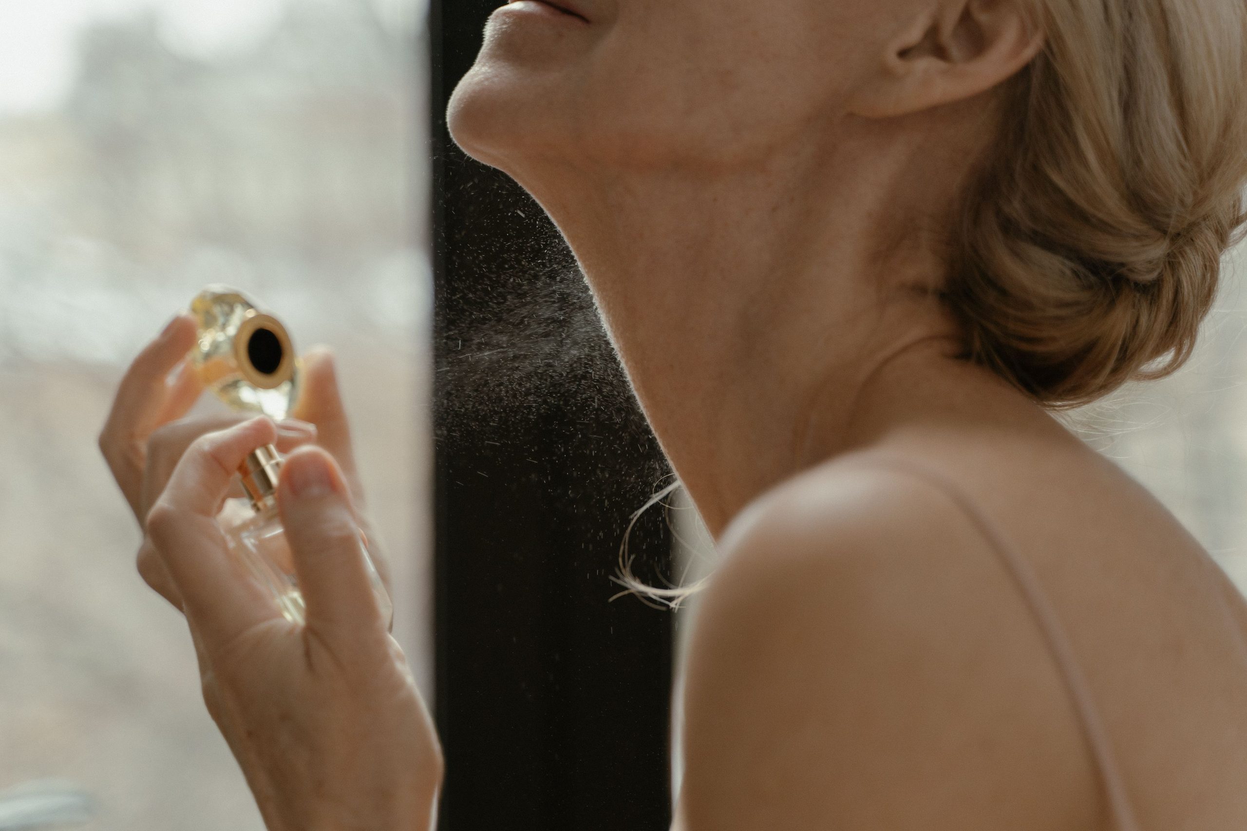 A woman applying perfume to her neck beside a bright window